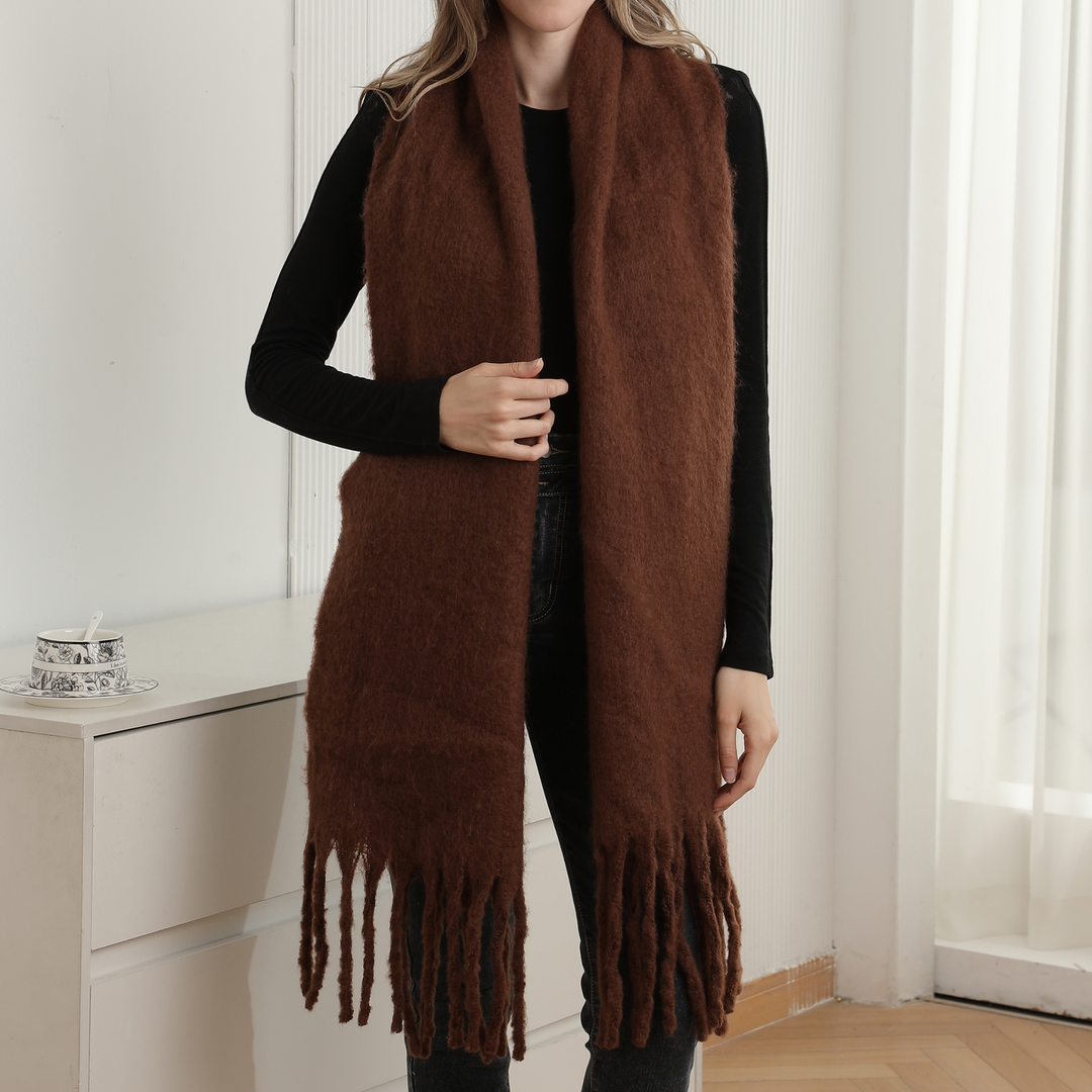 A woman wearing a long, fuzzy brown scarf with fringe ends over a black long-sleeve top and dark pants, standing indoors near a white cabinet and window.