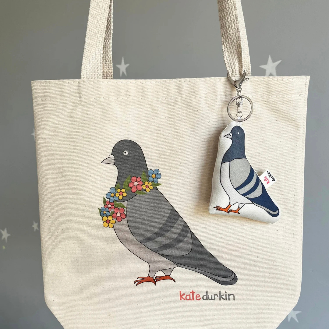 A fabric pigeon-shaped keychain charm with a silver lobster clasp and key ring, clipped to a tote bag with a design of a pigeon.