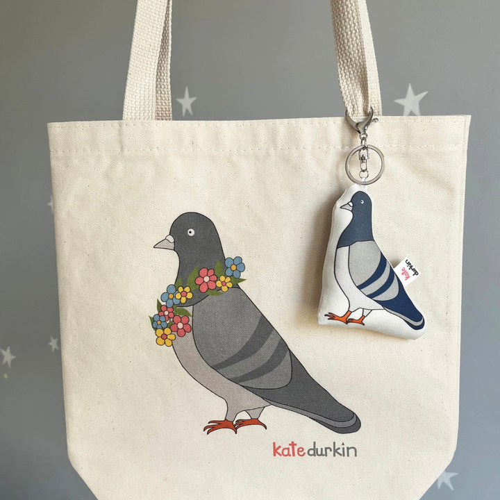 A fabric pigeon-shaped keychain charm with a silver lobster clasp and key ring, clipped to a tote bag with a design of a pigeon.
