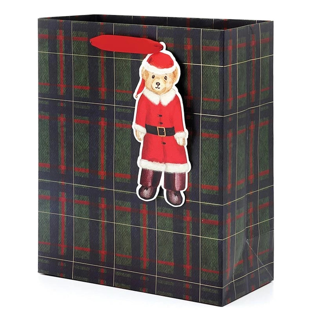 A Christmas-themed gift bag with a green and red plaid pattern, featuring a hanging die-cut ornament of a teddy bear dressed in a Santa suit and a red hanging loop.