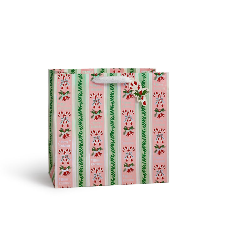 A small gift bag with a pink holiday pattern featuring candy canes, holly, and green pine branches, with white paper handles on a white background.