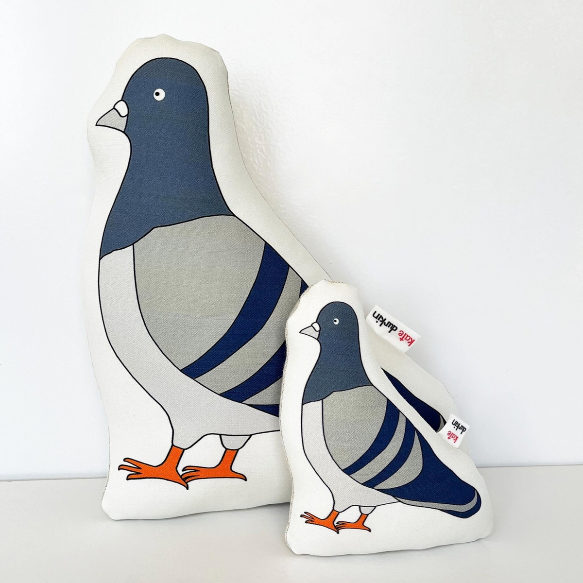 Two bird-shaped throw pillows in different sizes featuring a stylized pigeon design with a navy blue head, gray body, white wings, and orange feet, set against a plain white background.