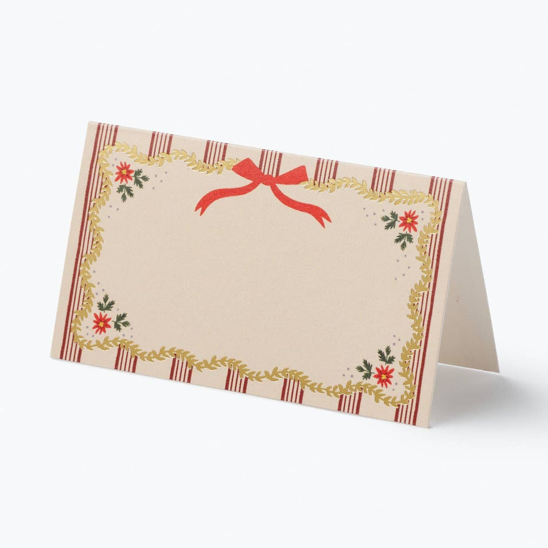 A small standing greeting card or place card with a cream center, a red ribbon bow at the top, and a decorative border with red stripes, gold leaf-like trim, and small red floral motifs, photographed on a light gray background.