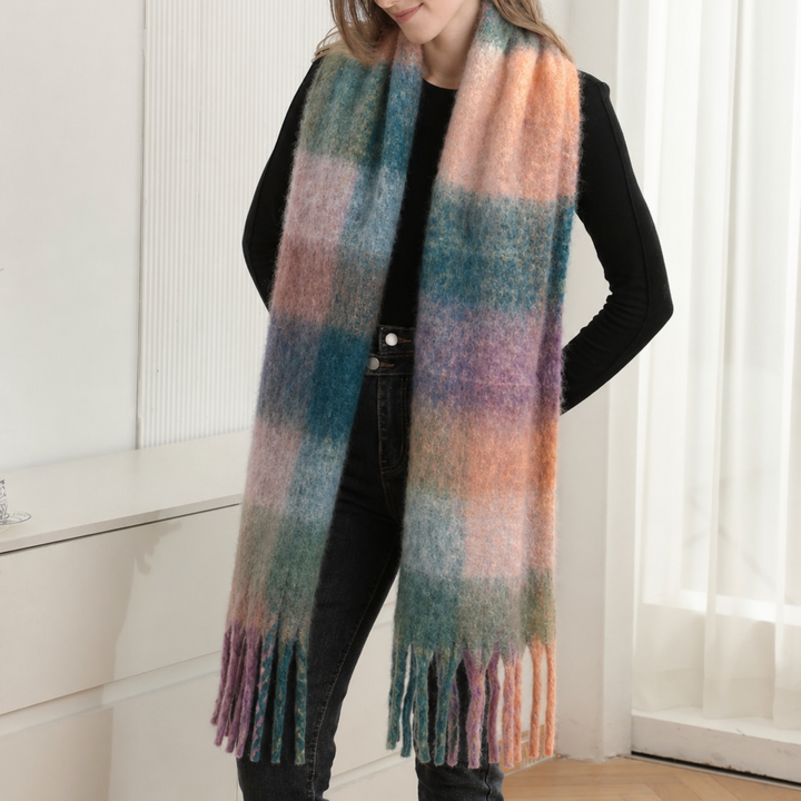 A woman wearing a long, fuzzy plaid scarf in soft pastel colors including blue, pink, peach, and purple, paired with a black top and dark jeans in a bright indoor setting.
