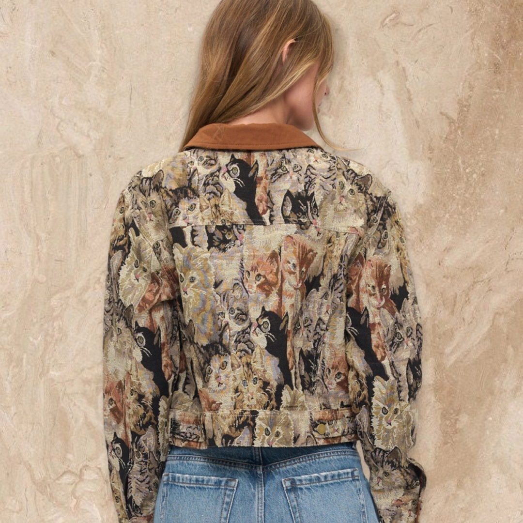 A woman viewed from behind wearing a long-sleeve cropped jacket with an all-over cat print and a brown collar, paired with blue jeans, standing against a textured beige wall.