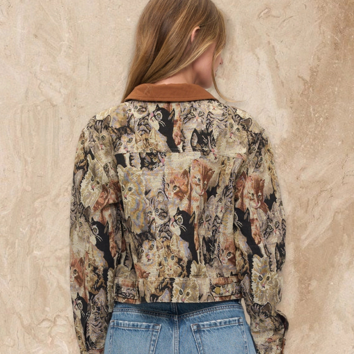 A woman viewed from behind wearing a long-sleeve cropped jacket with an all-over cat print and a brown collar, paired with blue jeans, standing against a textured beige wall.