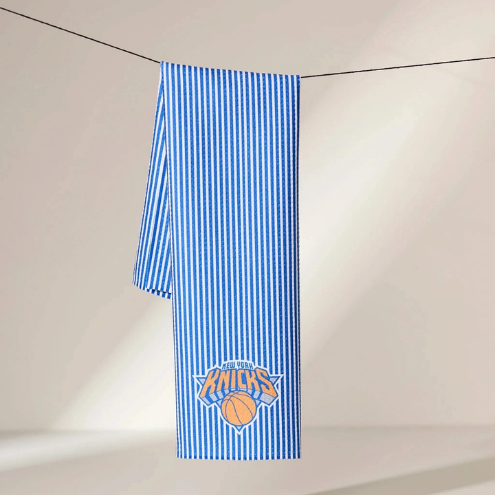 A blue and white vertically striped towel featuring the New York Knicks logo, hanging from a black string against a warm beige wall.