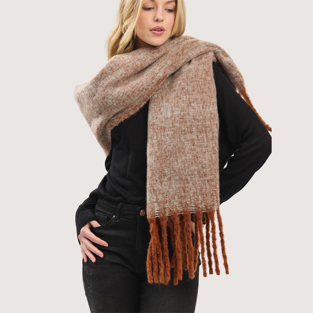 A woman wearing a thick, textured knit scarf in a heathered beige and brown color, styled over a black long-sleeve top and black jeans against a light gray studio background.