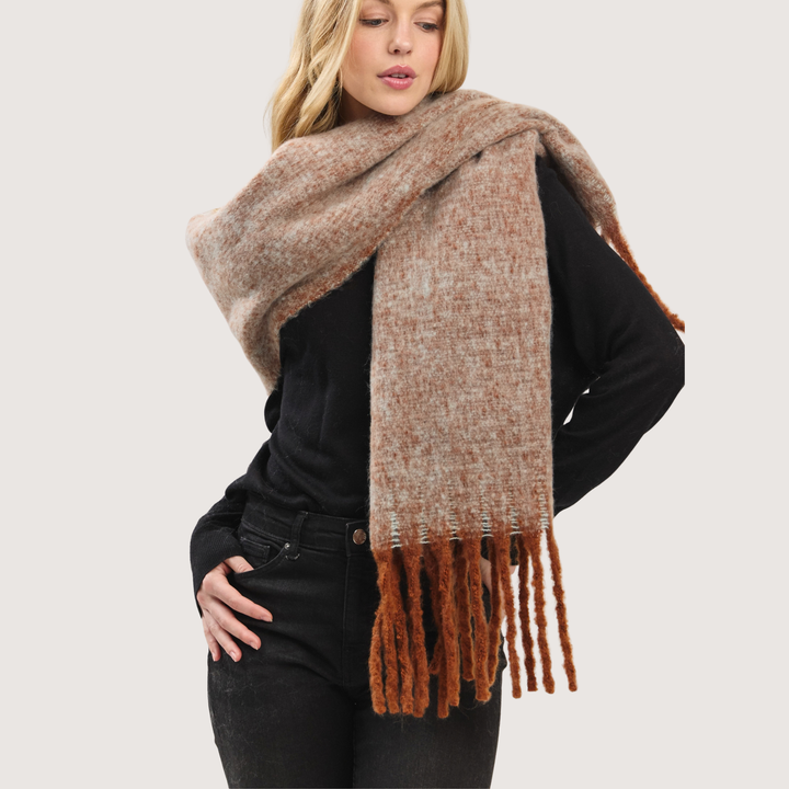 A woman wearing a thick, textured knit scarf in a heathered beige and brown color, styled over a black long-sleeve top and black jeans against a light gray studio background.