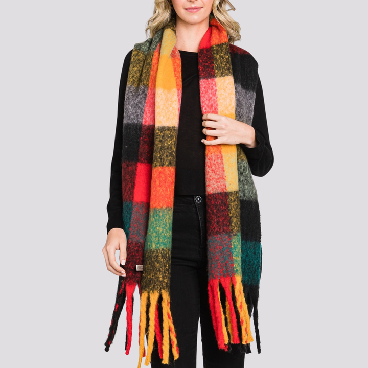 A woman wearing a long, multicolor plaid scarf with fringe ends over a black outfit, photographed against a plain light gray studio background.