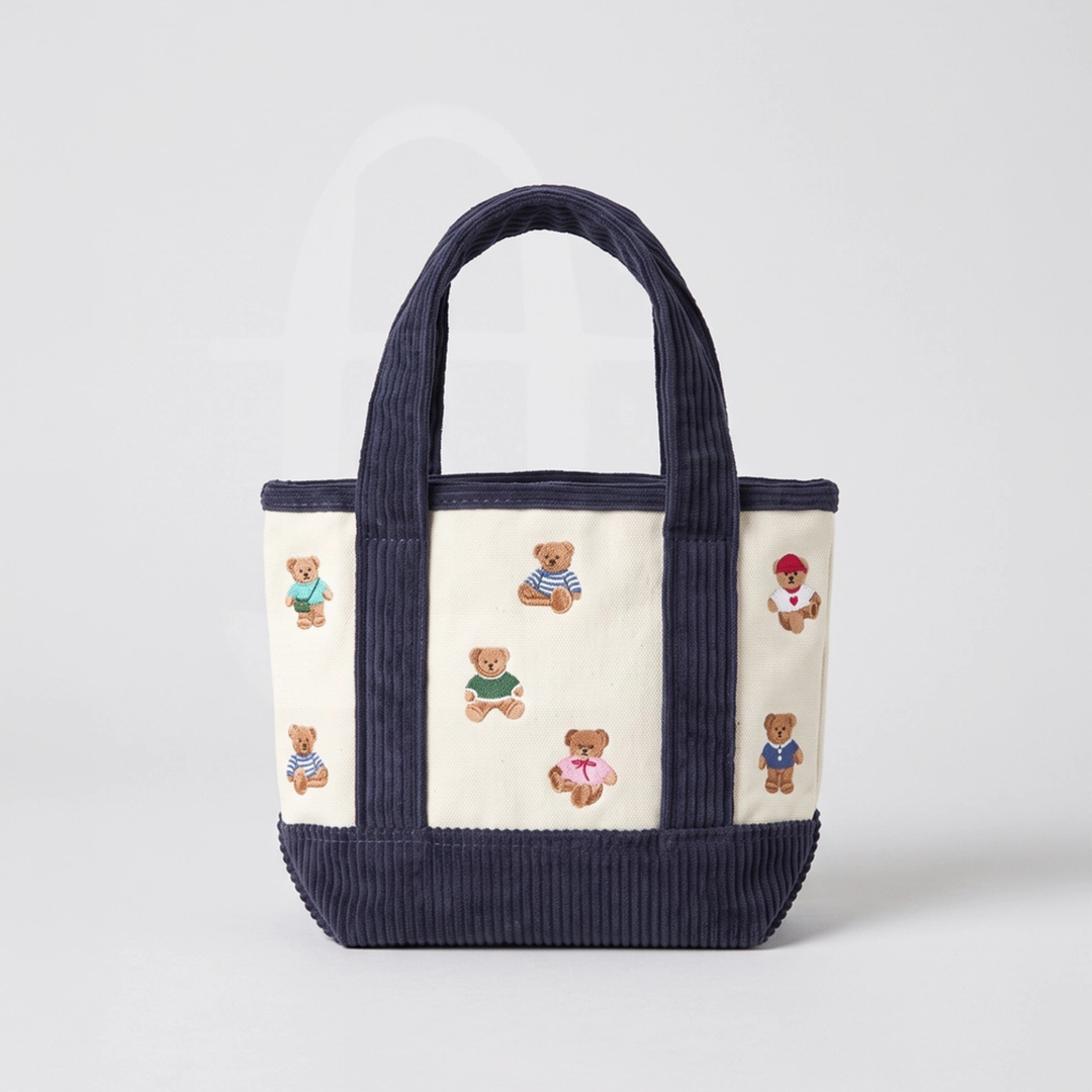 A cream-colored canvas tote bag with wide navy ribbed handles and a matching navy ribbed base, decorated with small embroidered teddy bears in various colorful outfits.