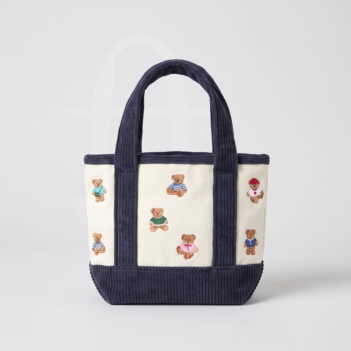 A cream-colored canvas tote bag with wide navy ribbed handles and a matching navy ribbed base, decorated with small embroidered teddy bears in various colorful outfits.