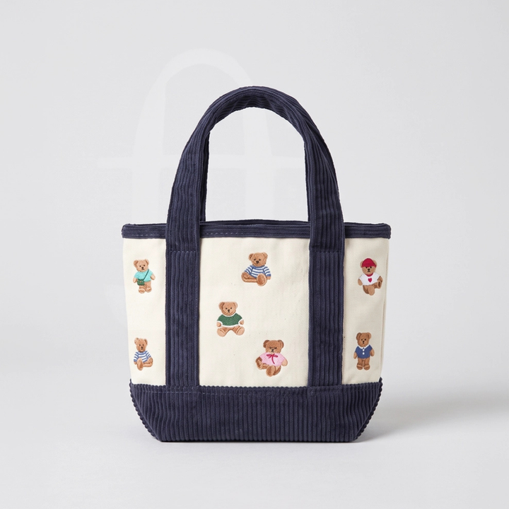 A cream-colored canvas tote bag with wide navy ribbed handles and a matching navy ribbed base, decorated with small embroidered teddy bears in various colorful outfits.