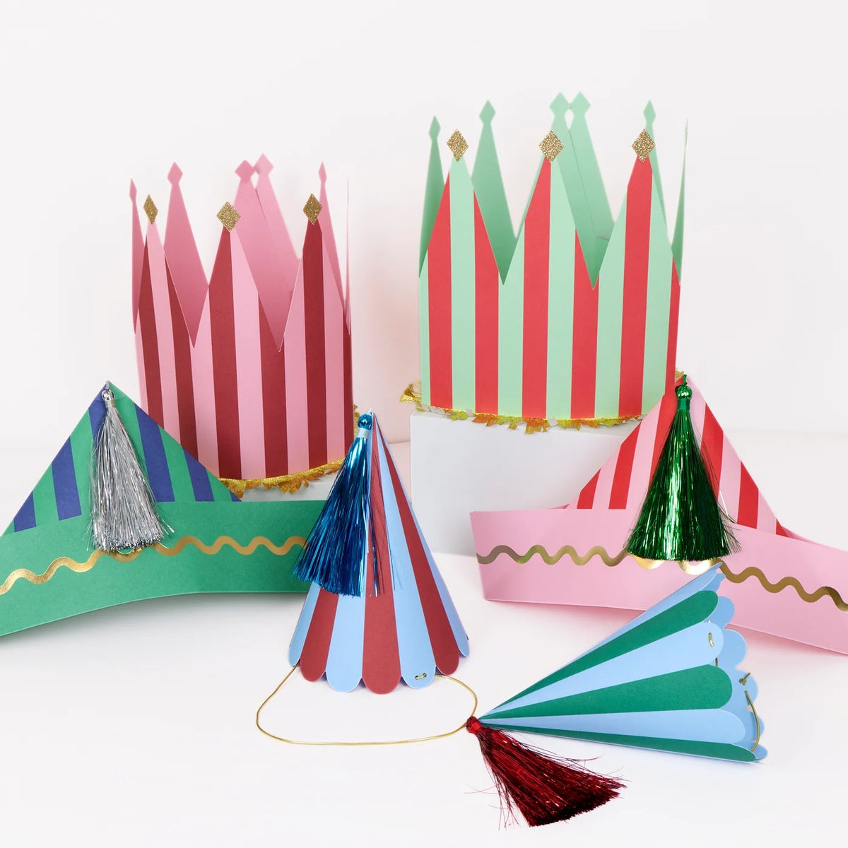 A set of colorful paper party hats and crowns with red, green, pink, and blue stripes, decorated with metallic gold trim and tassels, displayed on a clean white background.