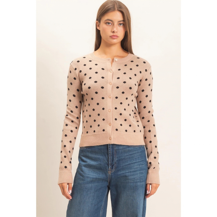 A woman modeling a beige cardigan with black polka dots and blue jeans against a plain off-white background.
