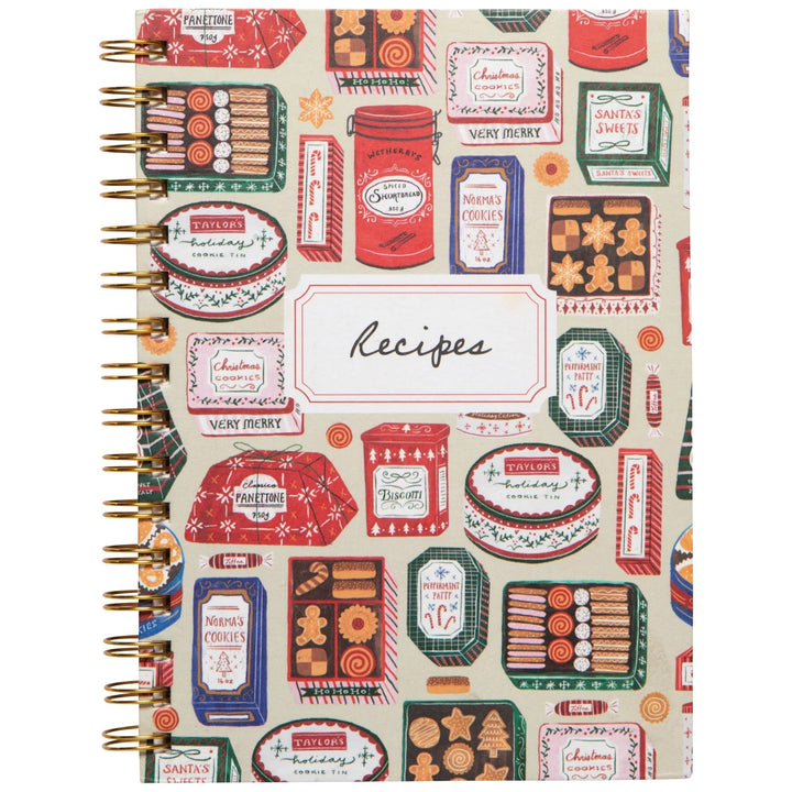 A spiral-bound recipe notebook with a vintage Christmas-themed cover pattern featuring gingerbread cookies, candy canes, and holiday boxes on a cream background. The center of the cover has a white label with the word &