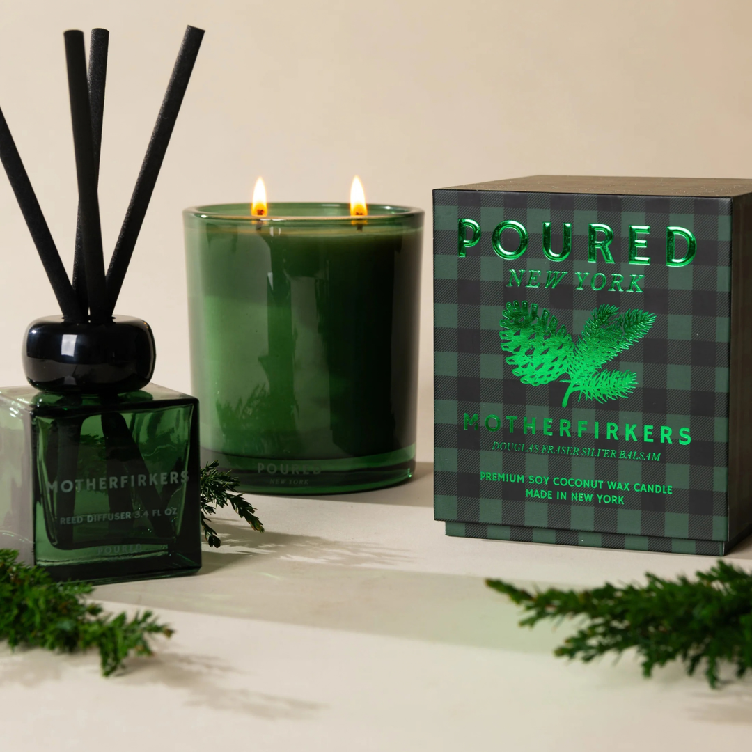 A green glass double-wick candle with two lit flames sits beside a matching green reed diffuser and its branded box. The box reads &