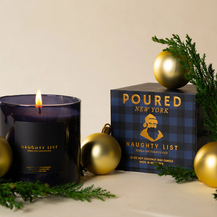 A dark purple glass candle with a lit flame sits beside its branded box labeled &