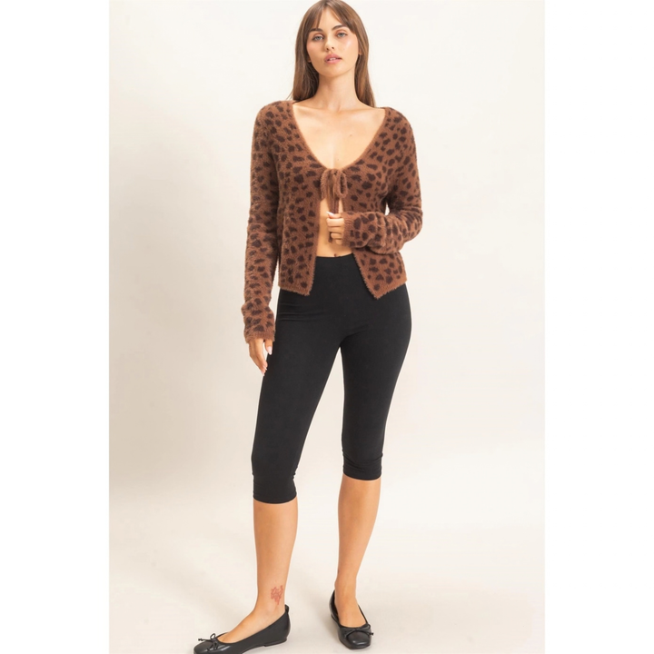 A woman modeling a brown leopard-print cardigan with a tie-front detail, paired with black capri leggings and black flats, against a plain light beige studio background.