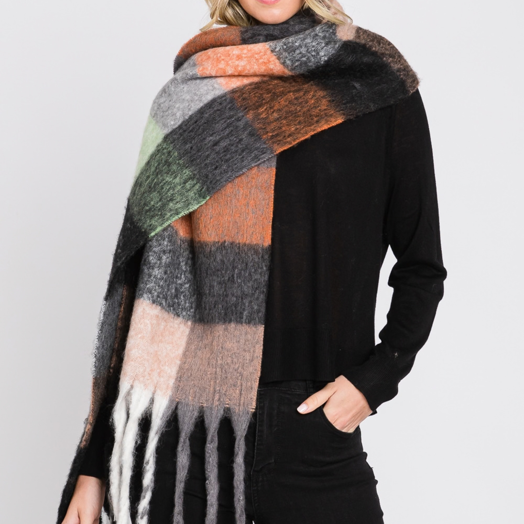 A woman wearing a large fuzzy plaid scarf in black, gray, orange, beige, green, and white, styled over a black long-sleeve top and black pants against a light gray studio background.