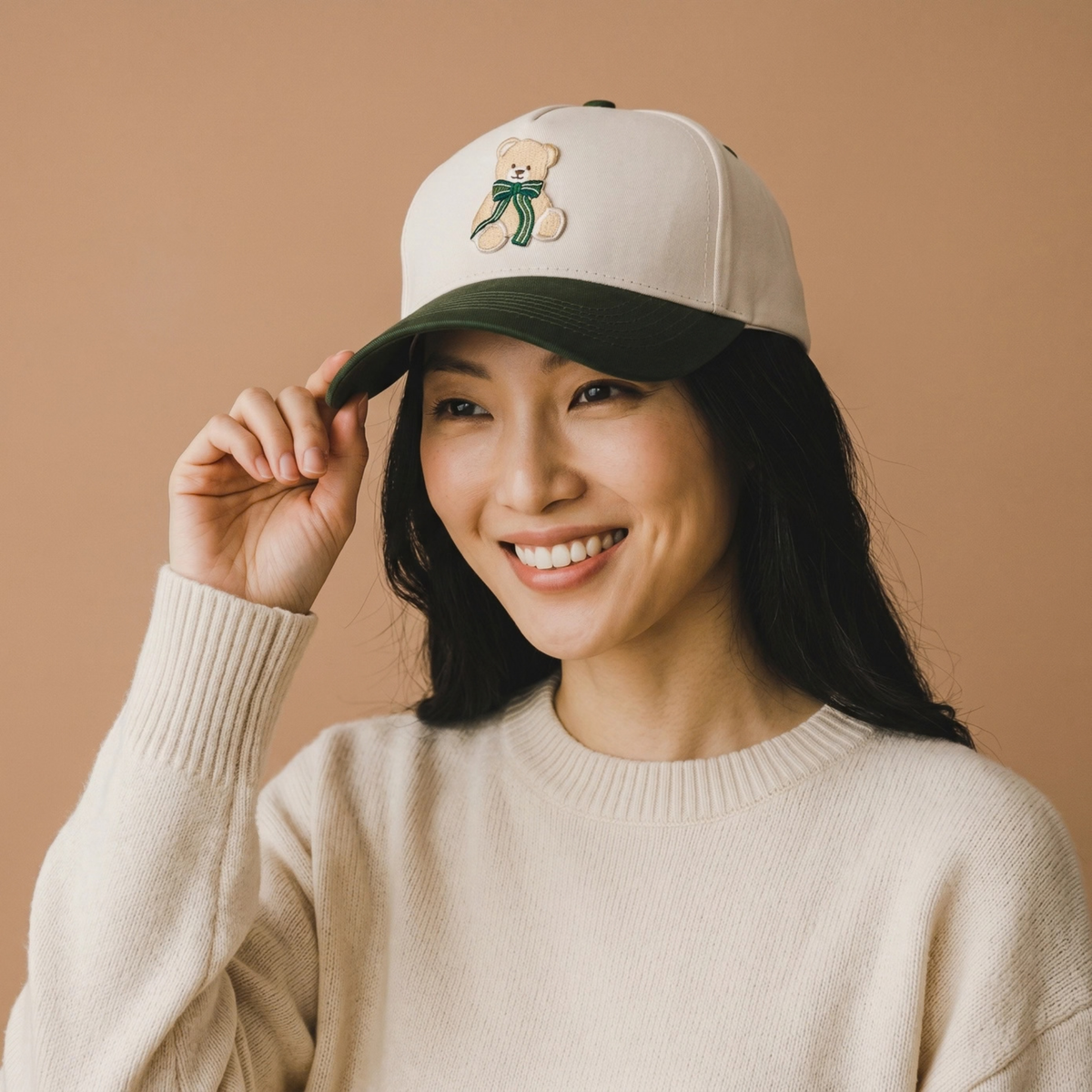 A woman smiling and adjusting a baseball cap with a cream crown, dark green brim, and an embroidered teddy bear with a green bow. She is wearing a cream knit sweater against a solid warm beige background.