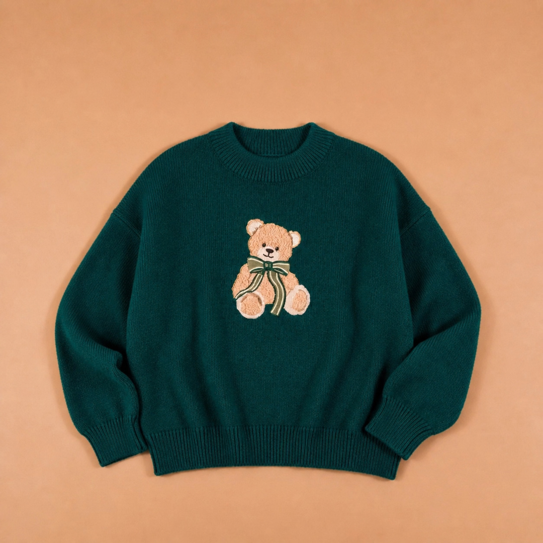 A dark teal green crewneck sweater featuring a knitted teddy bear motif on the chest, laid flat against a solid peach-colored background.