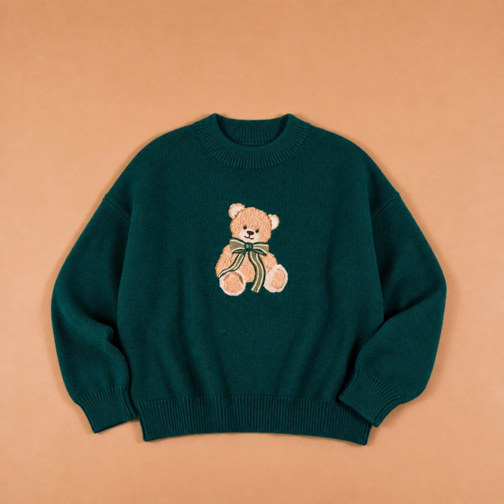 A dark teal green crewneck sweater featuring a knitted teddy bear motif on the chest, laid flat against a solid peach-colored background.