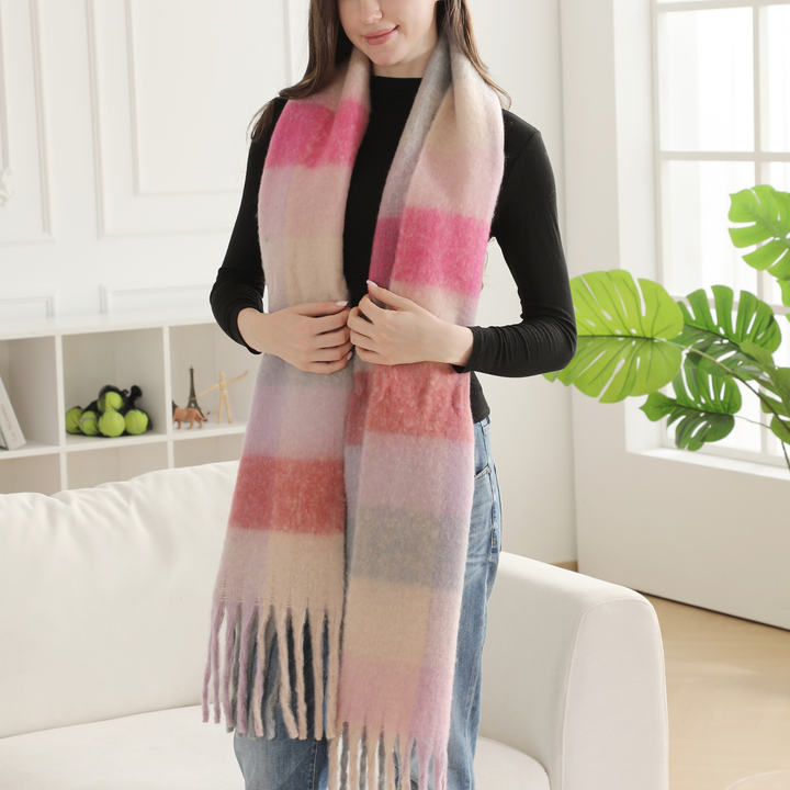 A woman wearing a long, soft plaid scarf in pink, beige, and light gray tones, styled over a black long-sleeve top and blue jeans in a bright, modern living room setting.