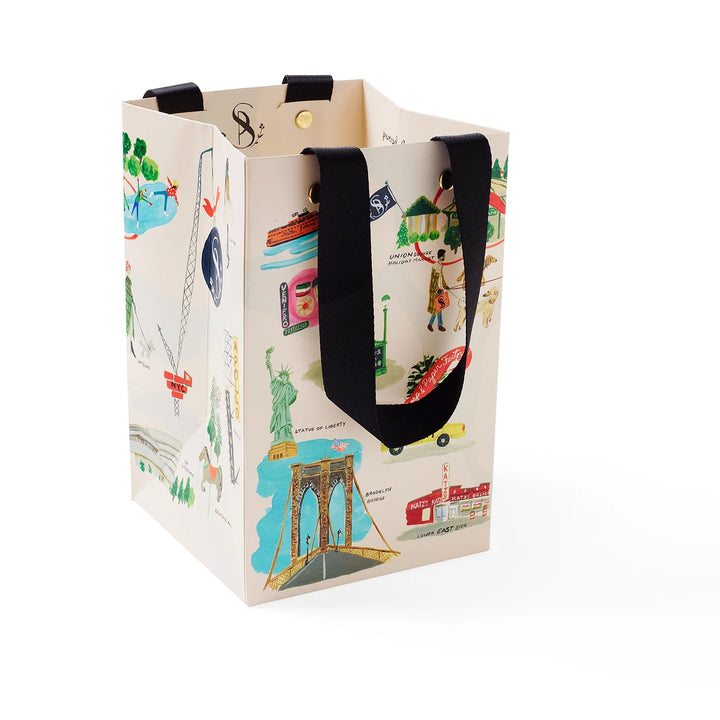 A cream-colored paper gift bag featuring a colorful travel-themed illustration print with black fabric handles, shown on a clean white background.