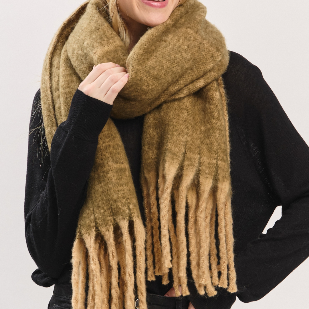 A close-up of a person wearing a thick, fuzzy mustard-brown fringed scarf over a black top, against a plain light gray background.