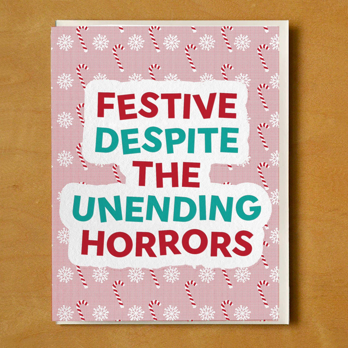 A square greeting card with a pink holiday pattern featuring white snowflakes and red-and-white candy canes, with the text 'FESTIVE DESPITE THE UNENDING HORRORS' in red and teal lettering on white torn-paper style blocks. The card is photographed flat against a warm brown background.