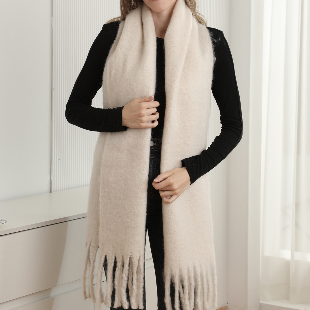 A woman wearing a long, soft cream-colored knit scarf with fringe ends over a black long-sleeve top, standing in a bright indoor setting.
