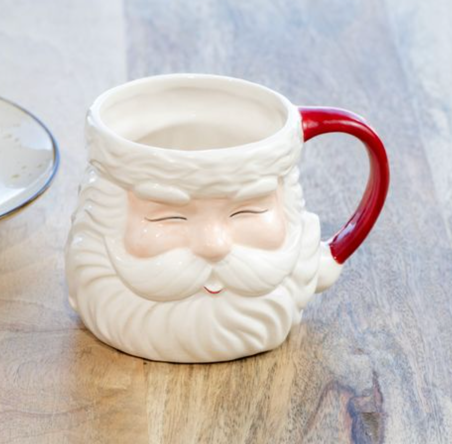 A ceramic mug shaped like Santa's face, with a white textured beard and hat, closed eyes, and a red handle, sitting on a wooden tabletop.
