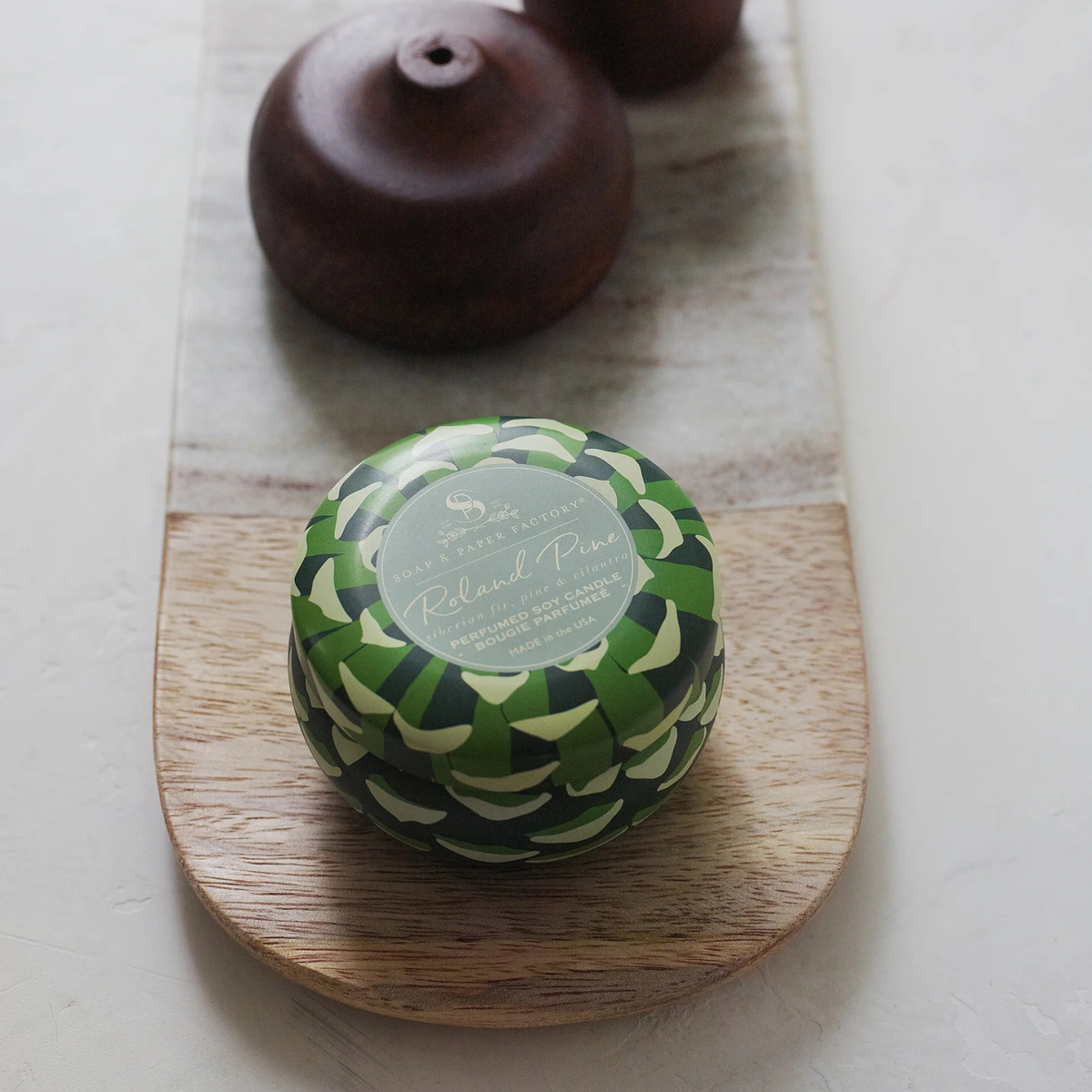 A round bar of soap wrapped in a green and black patterned paper sleeve sits on a wooden serving board, with two dark wooden objects behind it on a light surface.