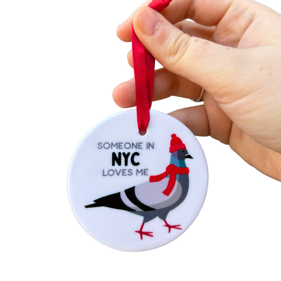 A hand holding a circular ornament that says "Some in NYC Loves Me," featuring a pigeon wearing a scarf and hat.