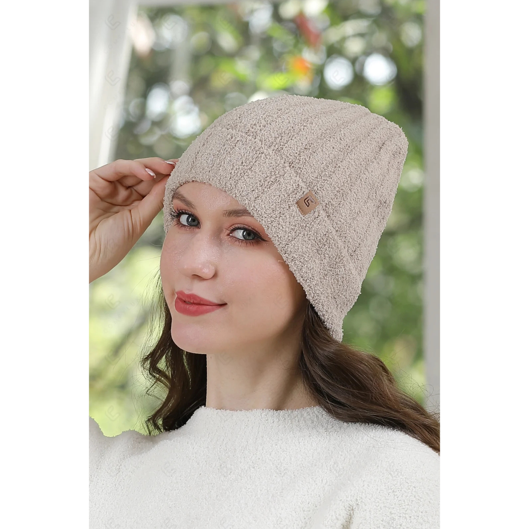 A woman wearing a beige textured knit beanie hat with a small brown logo tab, posed against a soft-focus outdoor background.