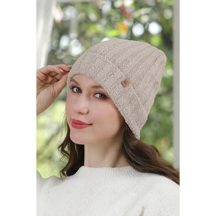 A woman wearing a beige textured knit beanie hat with a small brown logo tab, posed against a soft-focus outdoor background.