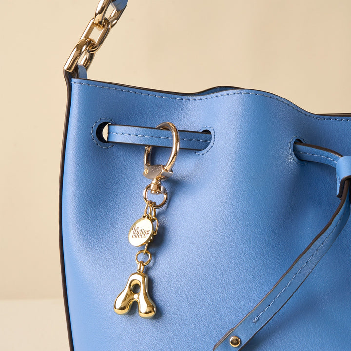 a blue handbag with a gold a shaped bag charm on it