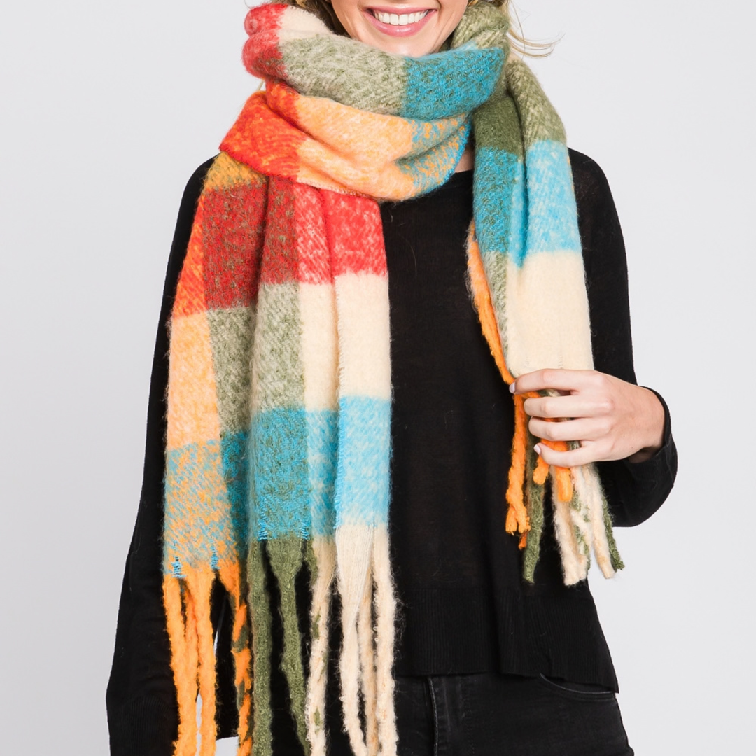 A person wearing a chunky knit scarf with large plaid blocks of orange, red, cream, light blue, olive green, and mustard yellow, paired with a black top against a light gray studio background.