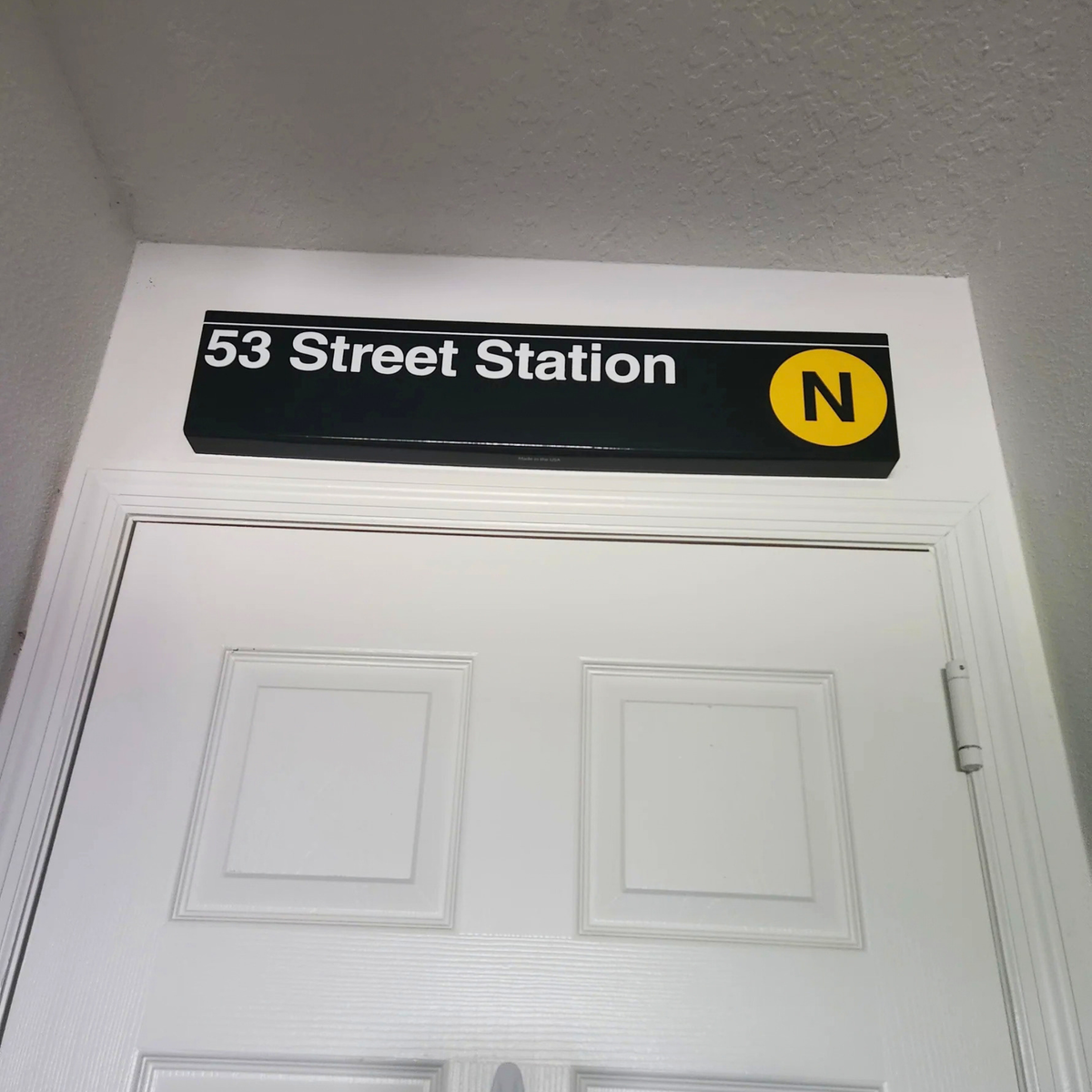 A black rectangular subway station sign reading '53 Street Station' with a yellow circle containing the letter N, mounted above a white door.