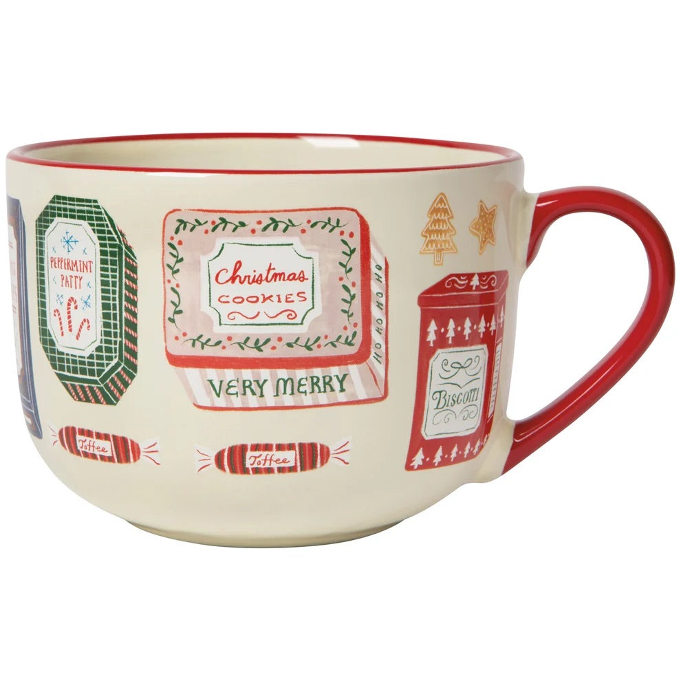 A ceramic Christmas mug with a cream-colored body, red handle and rim, decorated with festive labels reading 'Christmas Cookies', 'Very Merry', and other holiday designs.