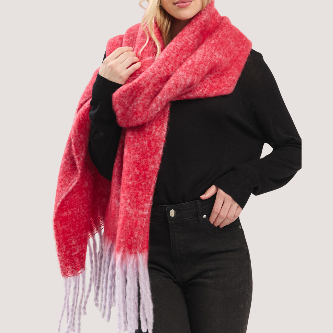 A woman wearing a chunky, fuzzy knit scarf in a coral-pink and light lavender gradient with fringed ends, styled over a black long-sleeve top and black jeans against a light gray studio background.