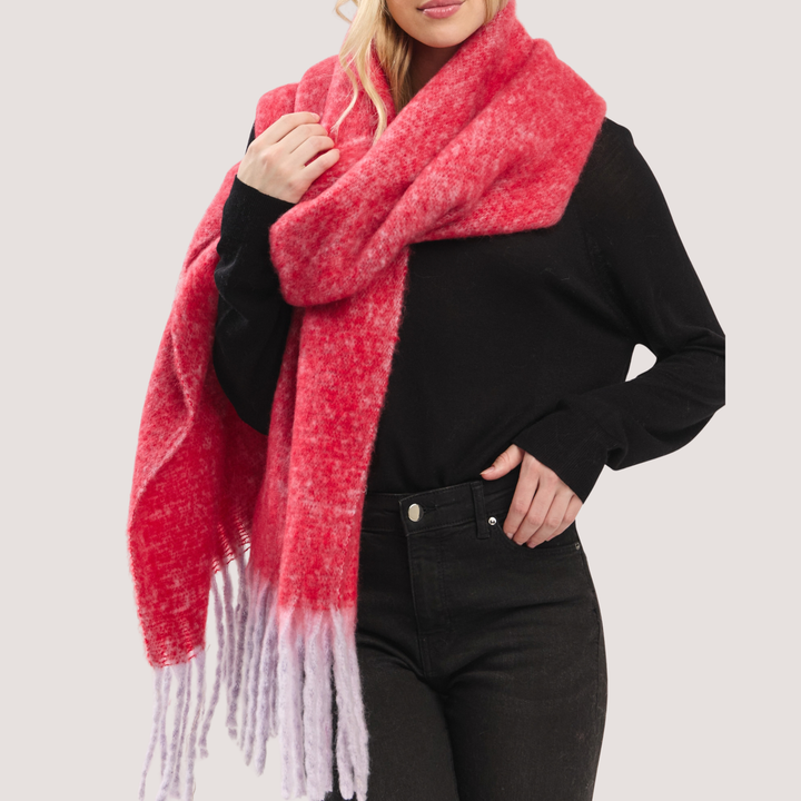 A woman wearing a chunky, fuzzy knit scarf in a coral-pink and light lavender gradient with fringed ends, styled over a black long-sleeve top and black jeans against a light gray studio background.