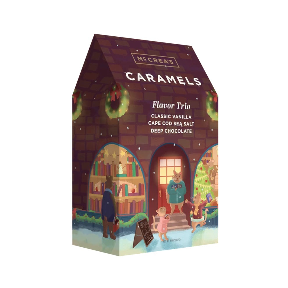 A house-shaped box of Micer's Caramels 'Flavor Trio' featuring holiday-themed illustrations of a cozy bookshop, a green wreath, and children on a snowy street, set against a plain white background.