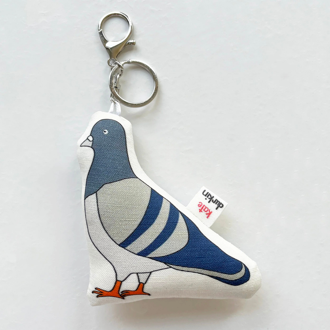 A fabric pigeon-shaped keychain charm with a silver lobster clasp and key ring, featuring a printed blue, gray, and white pigeon design with orange feet, on a light gray background.