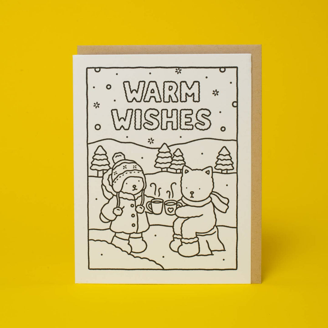 A square greeting card with a black-and-white winter illustration reading “WARM WISHES,” shown standing in front of a kraft envelope on a bright yellow background.