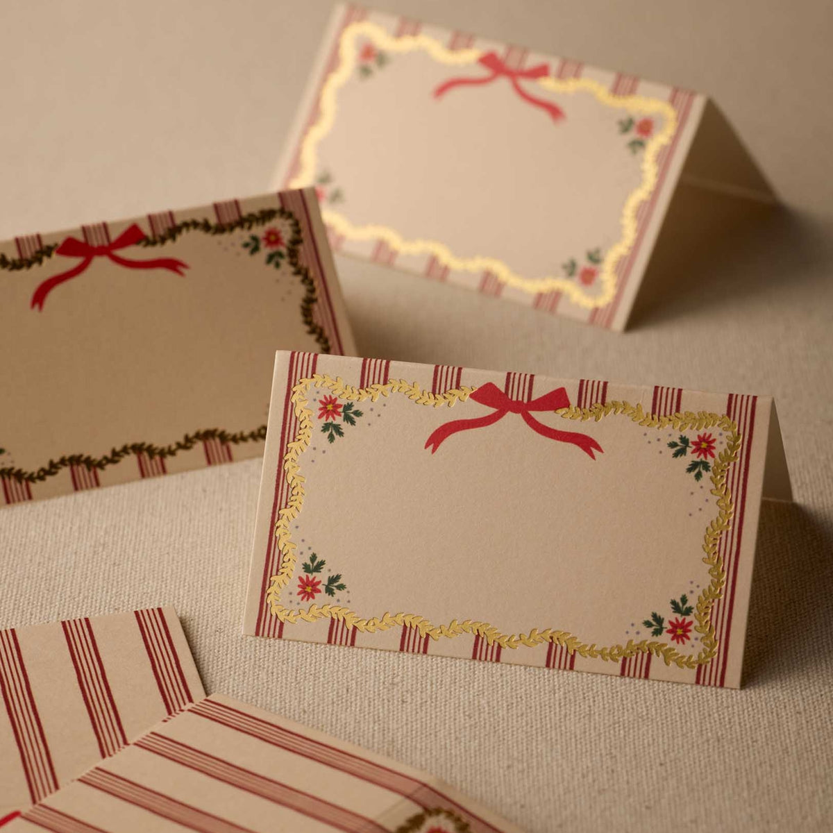 A set of decorative paper place cards with a vintage holiday design, featuring red ribbon bows, striped borders, and floral accents on a neutral textured background.