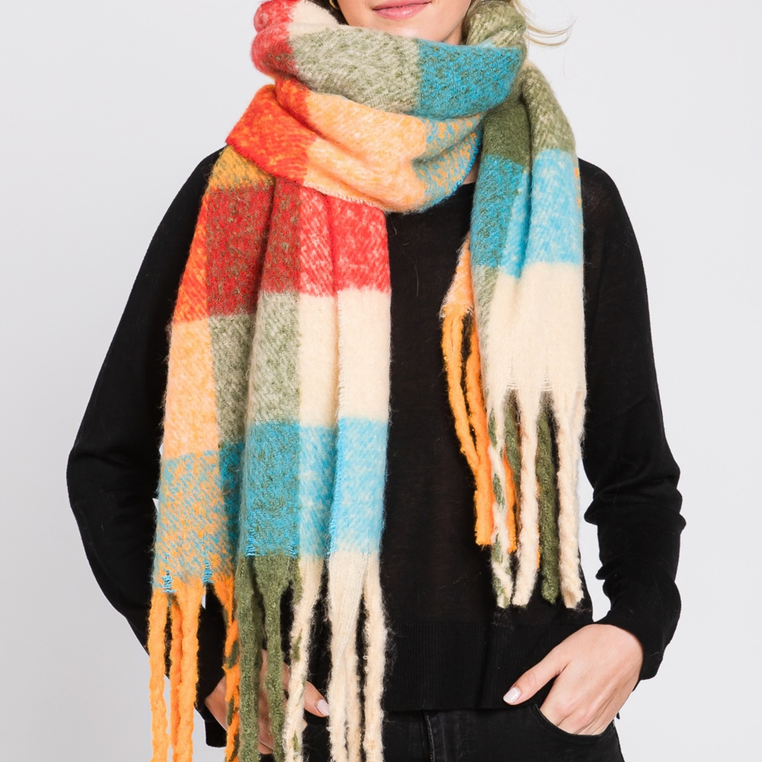 A person wearing a chunky, multicolor plaid scarf with fringe, styled over a black long-sleeve top against a light gray background.