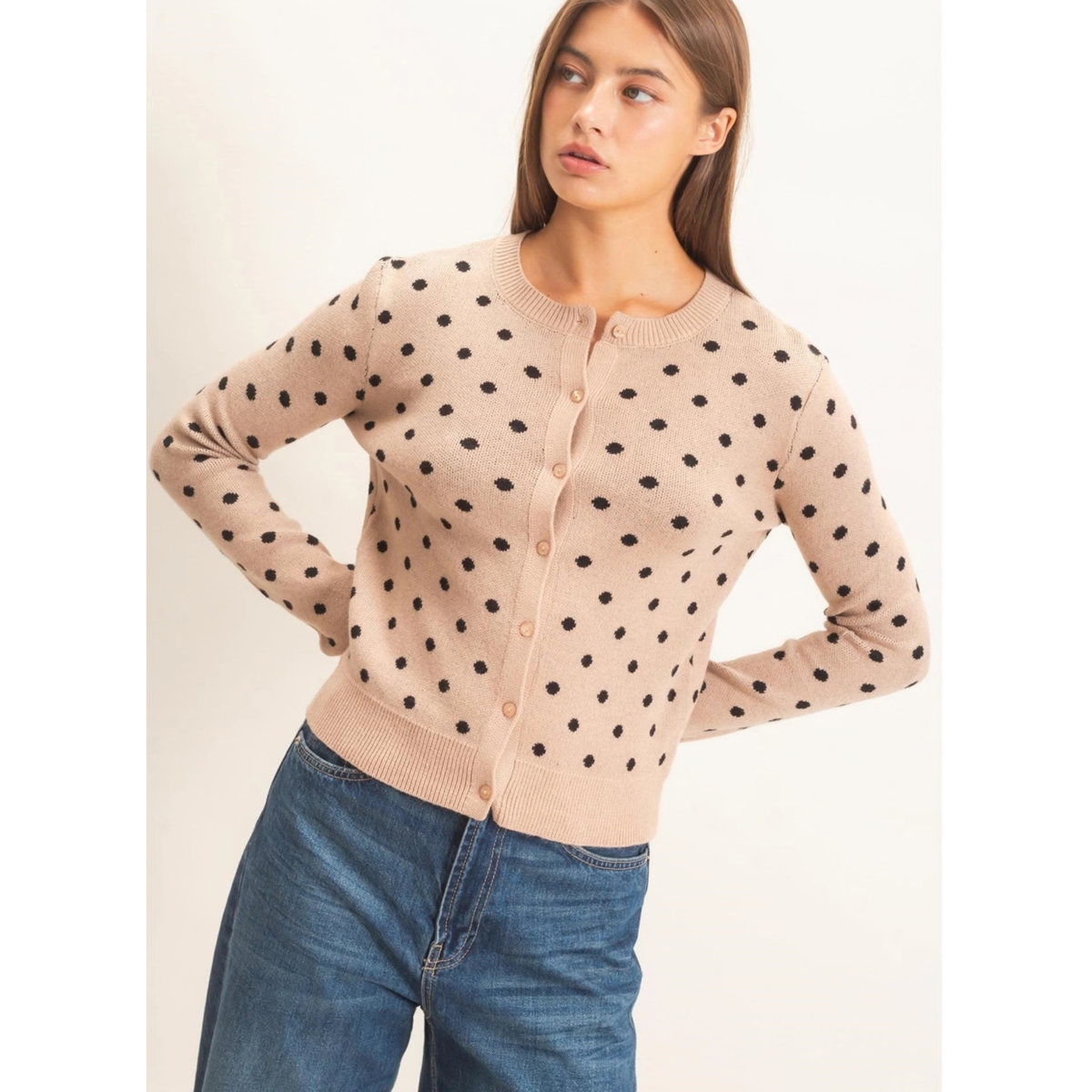 A woman wearing a beige cardigan with black polka dots and blue jeans, posing against a plain off-white background.