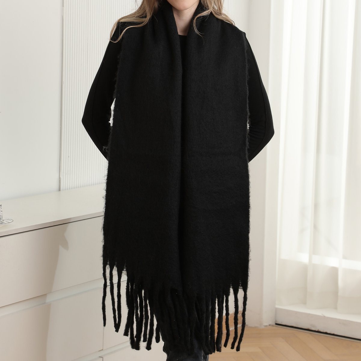 A person wearing a long black fuzzy wrap or scarf with fringe along the bottom, photographed indoors against a bright white wall and window curtains.